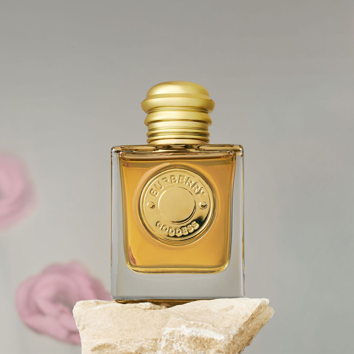 Quadratischer Glasflakon mit goldfarbenem Verschluss und Emblem, gefüllt mit bernsteinfarbenem Burberry Goddess Parfüm, das auf einem Steinblock ruht.