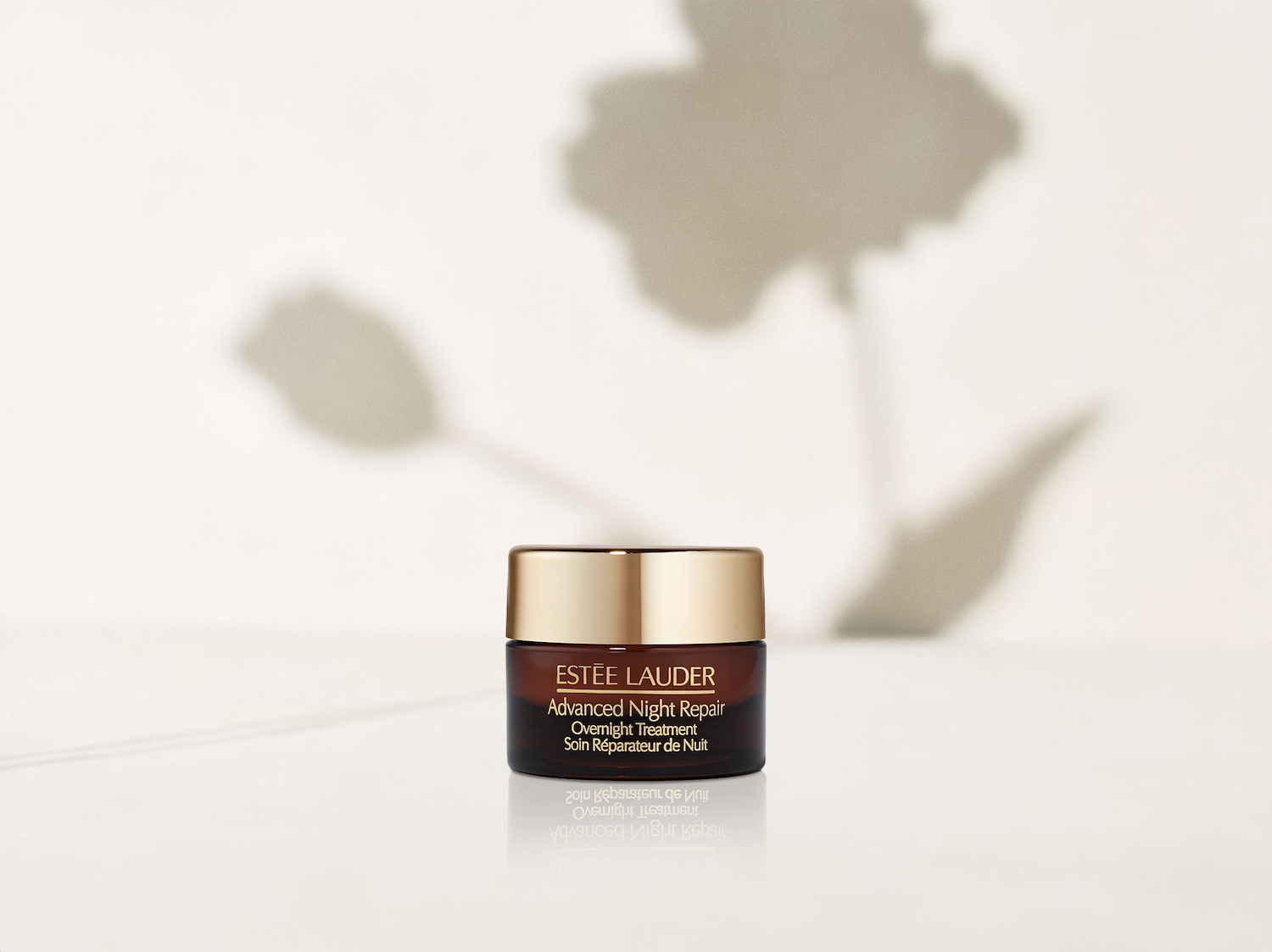 Eine Nahaufnahme eines Tiegel Estée Lauder Advanced Night Repair Overnight Treatment mit goldenem Deckel und braunem Tiegel, der auf einer glänzenden weißen Oberfläche steht.