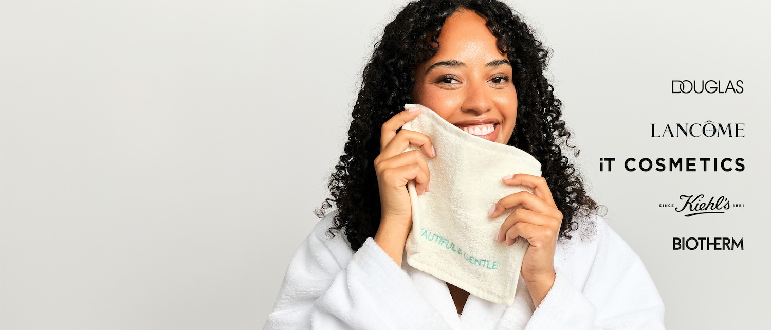Eine lächelnde Frau mit lockigem Haar hält ein cremefarbenes Handtuch mit der Aufschrift „BEAUTIFUL & GENTLE“ an ihr Gesicht, umgeben von den Logos von Douglas, Lancôme, IT Cosmetics, Kiehl's und Biotherm.
