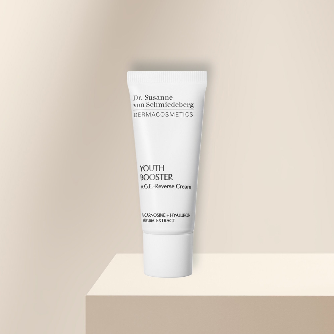 Die Dr. Susanne von Schmiedeberg Dermacosmetics Youth Booster A.G.E.-Reverse Cream in einer weißen Tube mit schwarzer Schrift, die L-Carnosine, Hyaluron und Yuyuba-Extrakt enthält, steht auf einem beigefarbenen Podest.