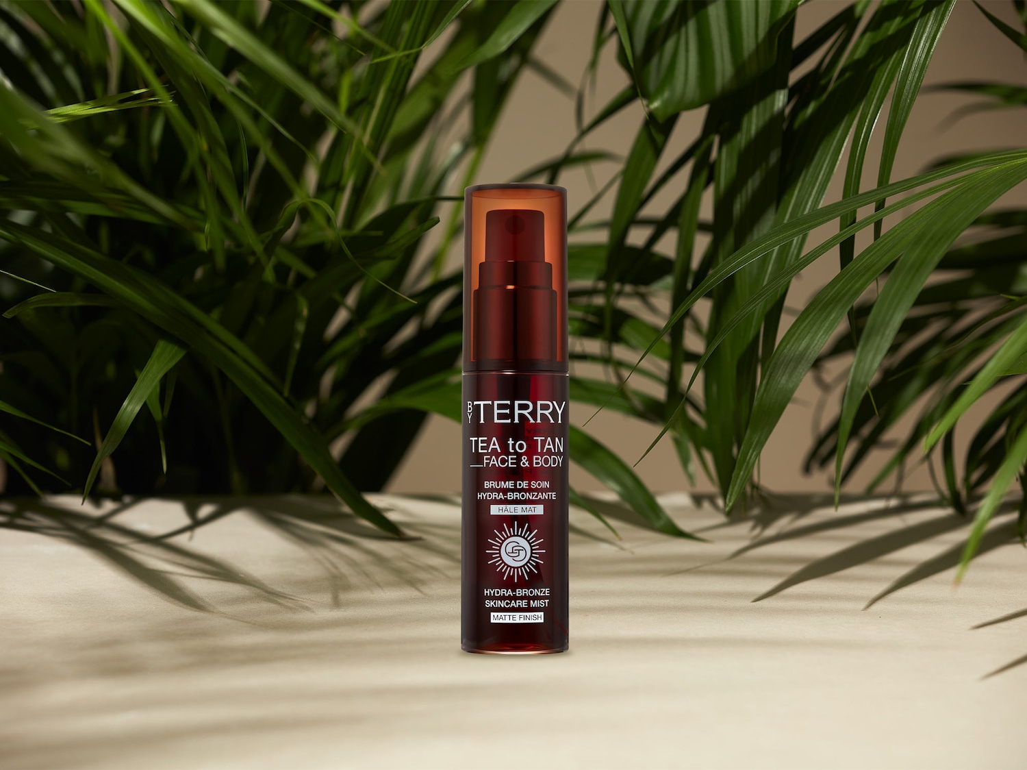 Das By Terry Tea to Tan Face & Body Hydra-Bronze Skincare Mist mit mattem Finish ist in einer braunen Sprühflasche mit orangefarbenem Deckel abgebildet und wird von grünen Blättern umrahmt.