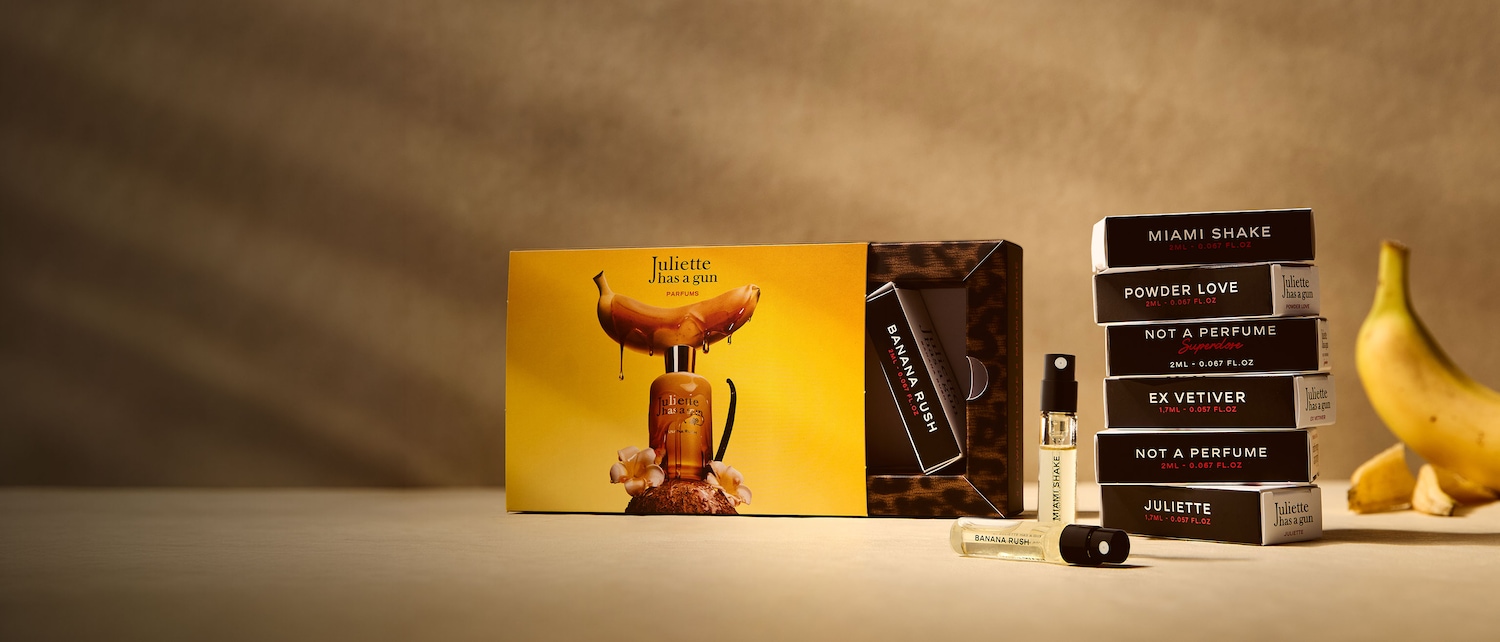 Das Juliette Has a Gun Parfümset präsentiert sich in einer gelben Verpackung mit einem Bananenmotiv, aus dem goldene Flüssigkeit tropft, umgeben von weißen Blüten und einer Vanilleschote.