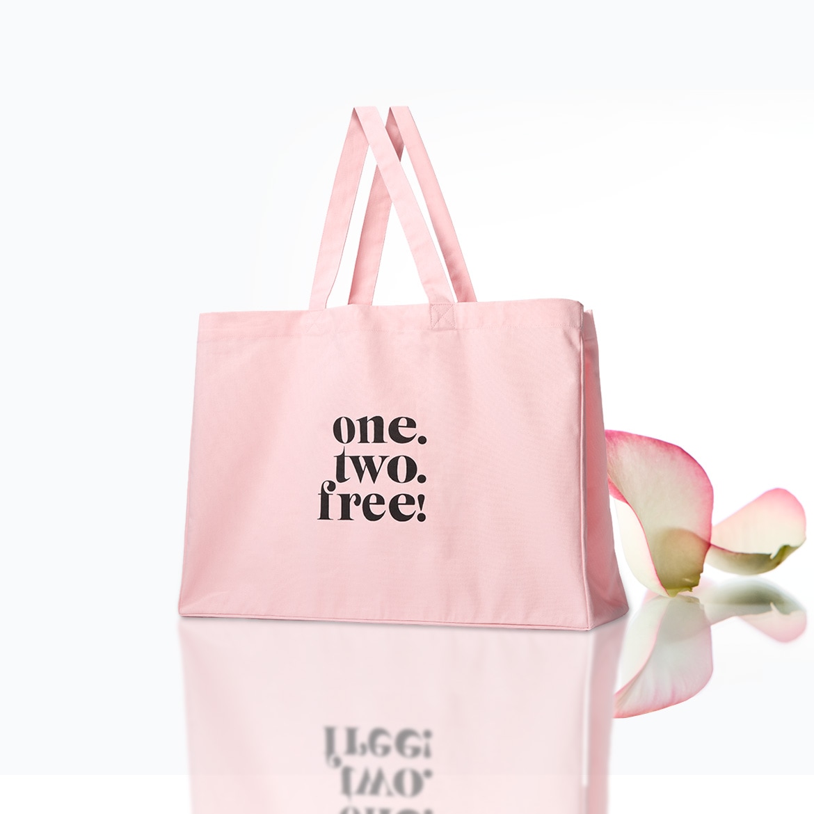 Eine hellrosa Stofftasche mit der Aufschrift „one. two. free!“ in schwarzer Schrift, daneben ein rosafarbenes Blütenblatt, das sich auf einer reflektierenden Oberfläche spiegelt.