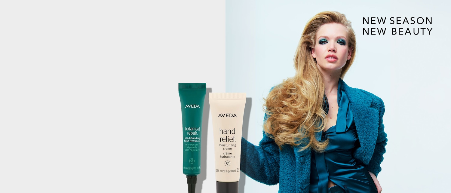 Eine Frau mit blonden Haaren und blauem Lidschatten trägt eine blaue Jacke und ein blaues Oberteil, daneben stehen zwei Tuben von Aveda-Produkten: Botanical Repair und Hand Relief.