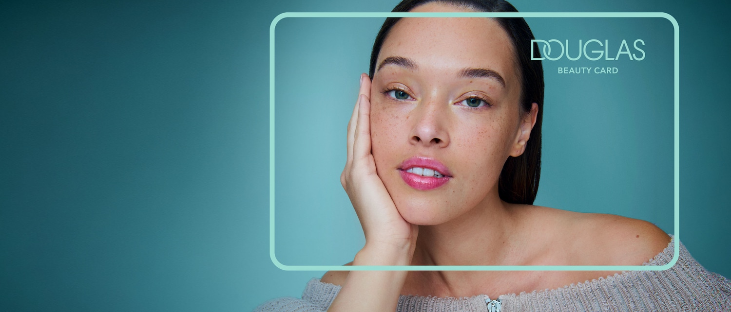 Eine Frau mit makelloser Haut und glänzenden rosa Lippen, die ihre Wange mit der Hand berührt, mit dem Douglas Beauty Card Logo in der oberen rechten Ecke.
