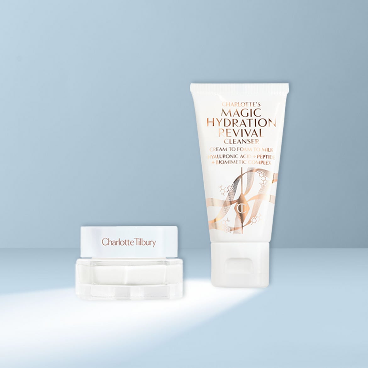 Charlotte Tilbury Magic Hydration Revival Cleanser, eine weiße Tube mit goldener Schrift, steht neben einem kleinen, durchsichtigen Tiegel mit weißem Deckel, auf dem „Charlotte Tilbury“ steht.