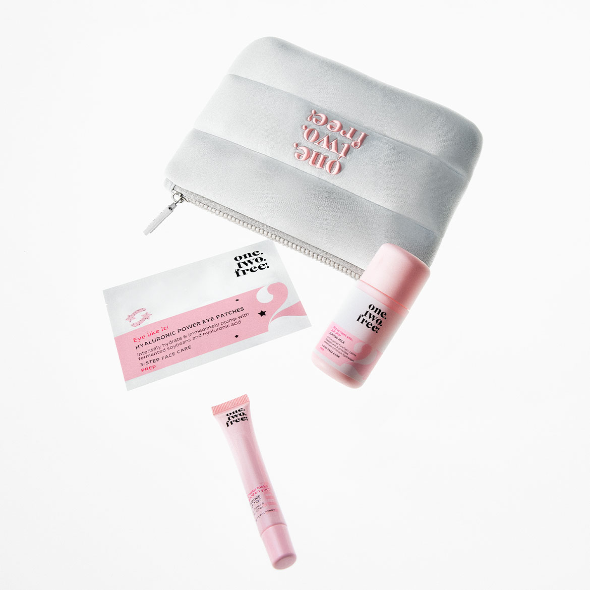 Das Bild zeigt ein "one.two.free!" Set, bestehend aus einer grauen Kosmetiktasche mit dem Logo, einer Packung Hyaluronic Power Eye Patches, einer Flasche Lazy Milk und einer Tube Lip Tint, alles in Rosa- und Weißtönen.