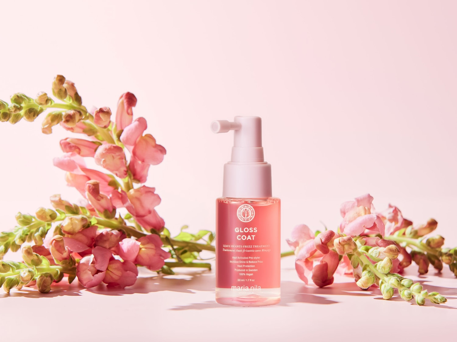 Das Maria Nila Gloss Coat Leave-In Anti-Frizz Treatment in einer 50-ml-Flasche mit rosa Flüssigkeit und weißem Pumpspender, umgeben von rosa Blüten, verspricht Glanz, weniger Frizz und Hitzeschutz.
