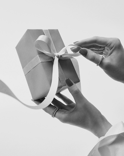 Eine Person öffnet eine hellgraue Geschenkbox, die mit einem weißen Band und einer Schleife verziert ist, ihre Hände sind mit filigranen Ringen geschmückt.
