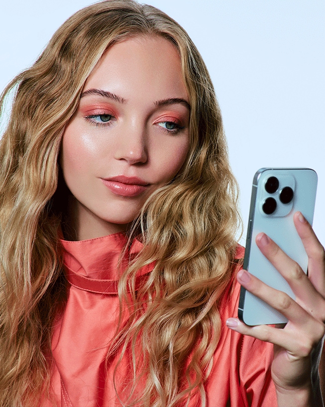 Eine junge Frau mit gewelltem, blondem Haar und einem korallfarbenen Oberteil macht ein Selfie mit ihrem Smartphone, wobei ihr passendes Augen-Make-up und ihre glänzenden Lippen im Fokus stehen