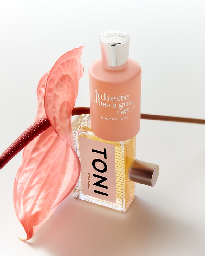 Zwei Parfümflaschen, eine in Pfirsichfarbe mit silbernem Verschluss und die andere klar mit einem goldenen Farbton, gestapelt mit einer rosa Anthurie-Blume daneben.